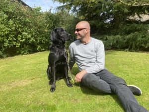 Photo shows myself and Spencer, my black lab, golden retriever cross, Guide dog. I am sat on the grass, with Spencer sat up beside me. I have a huge smile on my face as I look admiringly at my black beauty